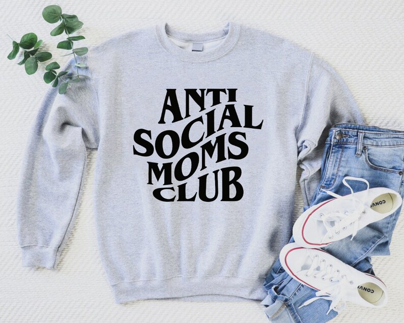 Puede incluir: Sudadera gris con texto negro que dice "Anti Social Moms Club" en una fuente retro. La sudadera est&aacute; colocada sobre una superficie blanca con un par de pantalones vaqueros azules y zapatillas blancas.
