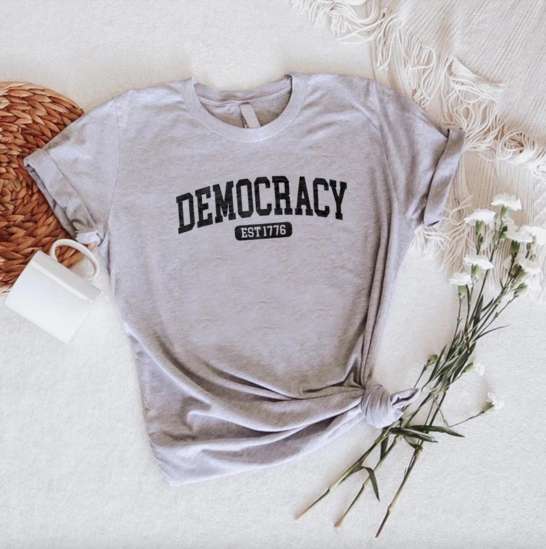 May include: A light gray t-shirt with the word "DEMOCRACY" in bold black letters arched across the chest, with "EST 1776" below. The shirt is knotted at the bottom and has rolled-up sleeves. A white mug and flowers are in the background.