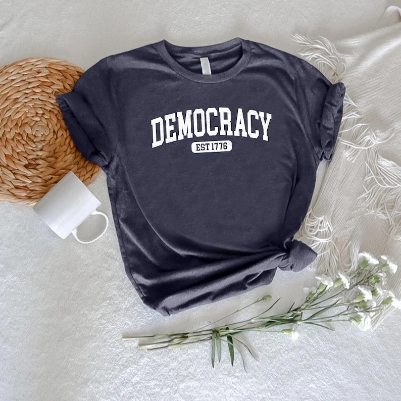 May include: A navy blue t-shirt with the word "DEMOCRACY" printed in white, arched across the top. Below the word "DEMOCRACY" is the text "EST 1776" in a smaller font. The shirt is knotted at the bottom.