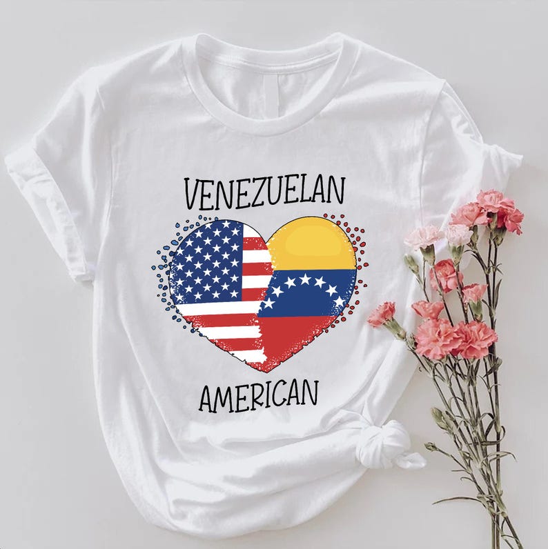 May include: White t-shirt featuring a heart-shaped design combining the American and Venezuelan flags. The words "VENEZUELAN" and "AMERICAN" are printed above and below the heart, respectively. A bouquet of pink flowers is to the right.