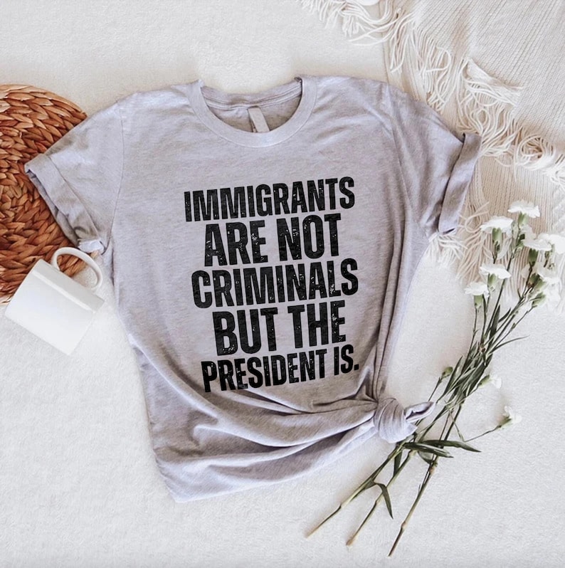 K&ouml;nnte beinhalten: Hellgraues T-Shirt mit dem Text "IMMIGRANTS ARE NOT CRIMINALS BUT THE PRESIDENT IS" in fetter schwarzer Schrift. Das T-Shirt ist am unteren Ende geknotet und liegt auf einer wei&szlig;en Oberfl&auml;che mit einer wei&szlig;en Tasse, einem geflochtenen Korb und wei&szlig;en Blumen.