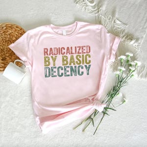 Puede incluir: Una camiseta rosa claro con el texto "RADICALIZED BY BASIC DECENCY" en letras rojas, doradas y turquesas. La camiseta est&aacute; anudada en la parte inferior. Tambi&eacute;n se ven una taza blanca, una cesta de mimbre y flores sobre una superficie blanca.
