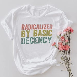 Puede incluir: Camiseta blanca con el texto "RADICALIZED BY BASIC DECENCY" en letras rojas, amarillas y turquesas. La camiseta tiene las mangas enrolladas y un nudo en el dobladillo. Flores rosas dispuestas a un lado.