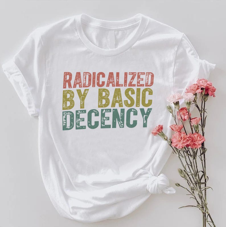 Puede incluir: Camiseta blanca con el texto "RADICALIZED BY BASIC DECENCY" en letras rojas, amarillas y turquesas. La camiseta tiene las mangas enrolladas y un nudo en el dobladillo. Flores rosas dispuestas a un lado.