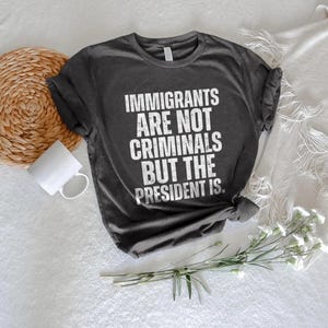 K&ouml;nnte beinhalten: Dunkelgraues T-Shirt mit dem wei&szlig;en Text "IMMIGRANTS ARE NOT CRIMINALS BUT THE PRESIDENT IS." Das Shirt liegt auf einer wei&szlig;en Oberfl&auml;che mit einem gewebten Untersetzer, einer wei&szlig;en Tasse und einem Zweig wei&szlig;er Blumen.