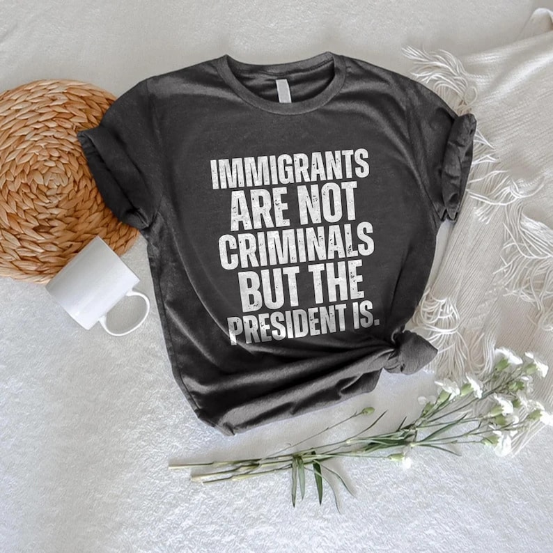K&ouml;nnte beinhalten: Dunkelgraues T-Shirt mit dem wei&szlig;en Text "IMMIGRANTS ARE NOT CRIMINALS BUT THE PRESIDENT IS." Das Shirt liegt auf einer wei&szlig;en Oberfl&auml;che mit einem gewebten Untersetzer, einer wei&szlig;en Tasse und einem Zweig wei&szlig;er Blumen.