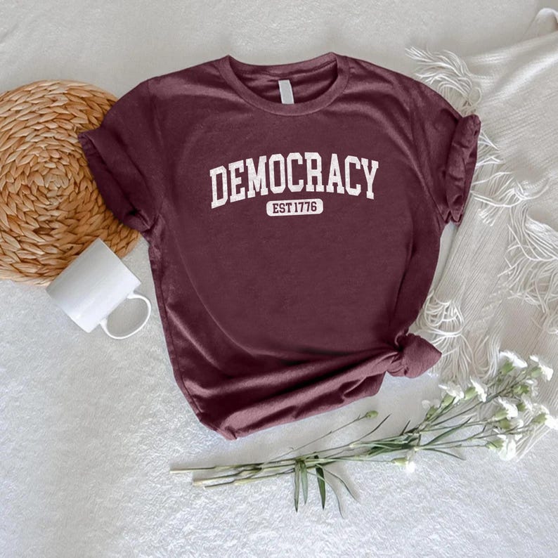May include: Maroon t-shirt with the word "DEMOCRACY" in white block letters and "EST 1776" below. The shirt is styled with rolled-up sleeves and a knot at the bottom. A white mug, woven mat, and flowers are in the background.