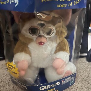 May include: A brown and white plush toy Gizmo from the movie Gremlins. The toy is in a clear plastic package with a blue and yellow background. The package says "Features the actual voice of Gizmo" and "Interactive Gizmo".