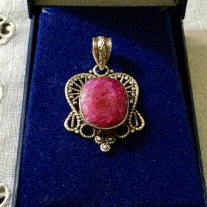Op de afbeelding: Een zilveren hanger met een grote, ovale, roze robijn die is gezet in een filigraanontwerp. De hanger bevindt zich in een blauwe fluwelen doos.