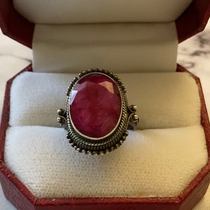 Op de afbeelding: Een zilveren ring met een grote, ovale robijn-edelsteen gezet in een decoratieve zilveren zetting. De ring bevindt zich in een rode fluwelen ringdoos.