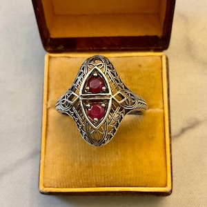 Peut inclure: Une bague en filigrane d'argent avec deux pierres de rubis rouges serties au centre. La bague est dans un écrin en velours jaune avec l'inscription "Au Roy d'Yvetot Rouen".