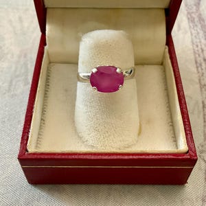 Peut inclure: Une bague en argent ornée d'une grosse pierre précieuse rose vif de forme ovale. La bague est présentée dans un écrin rouge avec un intérieur en velours blanc. Un bijou classique, idéal pour offrir.