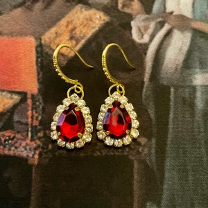 Peut inclure: Une paire de boucles d'oreilles pendantes dorées avec des cristaux rouges en forme de goutte d'eau entourés de petits cristaux transparents.