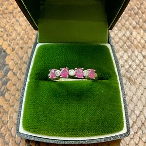 Pode incluir: Um anel com cinco pedras preciosas cor-de-rosa e pequenos diamantes brancos engastados em uma banda de metal branco. O anel é apresentado em uma caixa de anel de veludo verde com a inscrição "CHARLEROI" na tampa.