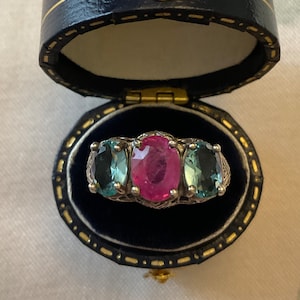 Op de afbeelding: Een zilveren ring met drie ovale edelstenen. De middelste steen is een roze robijn, en de twee ernaast zijn blauwgroene aquamarijnen.