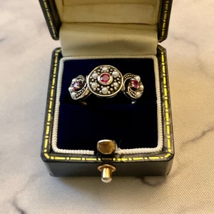 May include: A silver ring with three round settings, each with a red gemstone surrounded by white pearls. The ring is in a black velvet ring box.