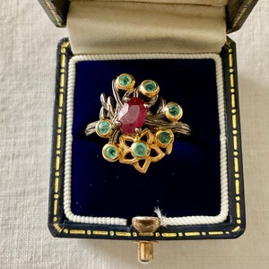 Peut inclure: Une bague avec une pierre rouge entourée de petites pierres vertes. La bague est sertie d'argent et d'or et présente un motif floral.