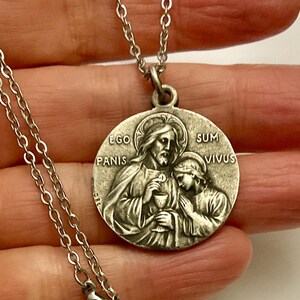 Peut inclure: Collier pendentif argenté avec un médaillon religieux circulaire. Le médaillon représente deux figures, l'une tenant un calice, avec l'inscription latine "EGO PANIS SUM VIVUS". La chaîne est de type maillon fin.