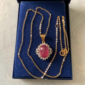 Könnte beinhalten: Eine goldene Kettenkette mit einem rosa ovalen Edelsteinanhänger, der von kleineren klaren Edelsteinen umgeben ist. Die Kette befindet sich in einer blauen Samtbox.