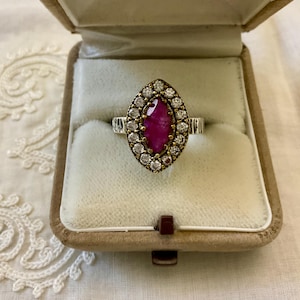 Könnte beinhalten: Ein Vintage-Ring mit einem markisenförmigen Rubin-Mittelstein, umgeben von kleinen, runden Diamanten. Der Ring ist in einem Gold- und Silberband gefasst und wird in einer samtgefütterten Schmuckschatulle präsentiert.