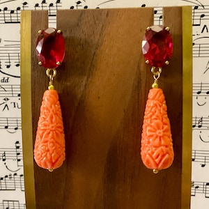Peut inclure: Une paire de boucles d'oreilles pendantes avec une pierre précieuse rouge facetée et une goutte de corail sculptée. Le corail est de couleur orange clair et présente un motif floral.