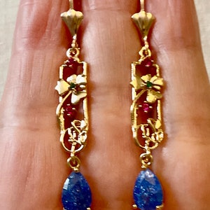 Peut inclure: Une paire de boucles d'oreilles pendantes en or avec une pierre bleue en forme de larme au bas. Les boucles d'oreilles présentent un délicat motif floral avec des accents rouges et verts.