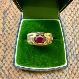 Peut inclure: Une bague en or avec une grosse pierre centrale en rubis de forme ovale. La bague est sertie dans un anneau en or avec de plus petites pierres précieuses de chaque côté du rubis. La bague est présentée dans un écrin vert doublé de velours.