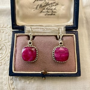 Op de afbeelding: Zilveren oorbellen met grote, vierkante, robijnrode edelstenen. De oorbellen worden gepresenteerd in een vintage blauwe sieradendoos met de tekst "Jewellers Estd. 1692. A. Woodhouse & Son Ltd."