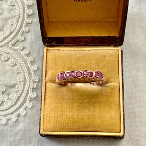 Peut inclure: Une bague en or ornée de cinq rubis ronds rose foncé alignés, présentée à l'intérieur d'un écrin à bijoux carré de style vintage, doté d'une doublure en velours jaune moutarde et d'un cadre extérieur marron foncé.