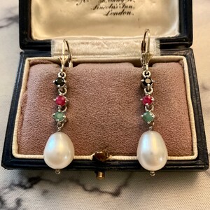 Peut inclure: Une paire de boucles d'oreilles en or avec une perle pendante et une chaîne de pierres précieuses noires, rouges et vertes. Les boucles d'oreilles sont dans une boîte doublée de velours.