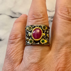 Peut inclure: Une bague large en argent avec une grande pierre précieuse rose entourée de petites pierres précieuses vertes et claires. La bague a une surface texturée et une bordure dorée autour de la grande pierre précieuse.