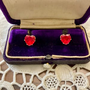 Peut inclure: Une paire de boucles d'oreilles en forme de cœur avec des pierres précieuses rouges serties d'or. Les boucles d'oreilles sont dans un écrin à bijoux en velours violet.