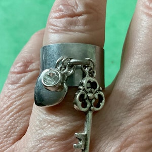 Peut inclure: Une bague en argent avec un charme en forme de cœur et une clé. La bague est large et a une surface lisse. Les charmes sont attachés à la bague par une chaîne.