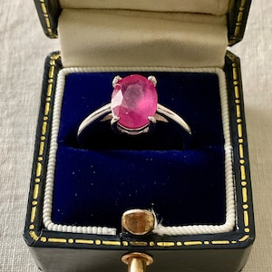 Peut inclure: Une bague en argent avec une pierre précieuse ovale en rubis rose, présentée dans un écrin à anneaux en velours bleu.