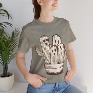 Puede incluir: Una camiseta gris con un gráfico marrón de un grupo de cactus con caras. Los cactus están en una maceta marrón.