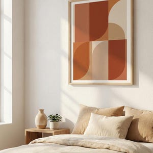 May include: A framed abstract art print with geometric shapes in shades of orange, brown, and beige hangs above a bed with beige bedding and pillows. A small wooden side table holds a vase and a potted plant.