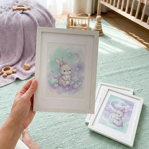 May include: White framed art prints featuring watercolor illustrations of a bunny with stars and a moon. The bunny is sitting on stars in a dreamy, pastel-colored night sky. The prints are displayed in a nursery setting.