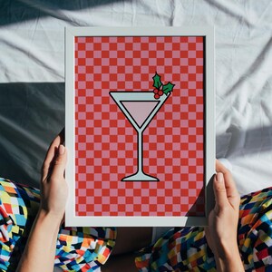 May include: A framed art print depicting a pink cocktail with a holly garnish set against a red and pink chequered backdrop. The cocktail glass is outlined in black and filled with pink liquid. The frame is white.
