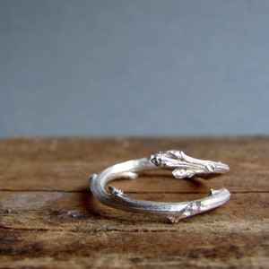 May include: A silver ring shaped like a twisted branch with small details. The ring is on a wooden surface.