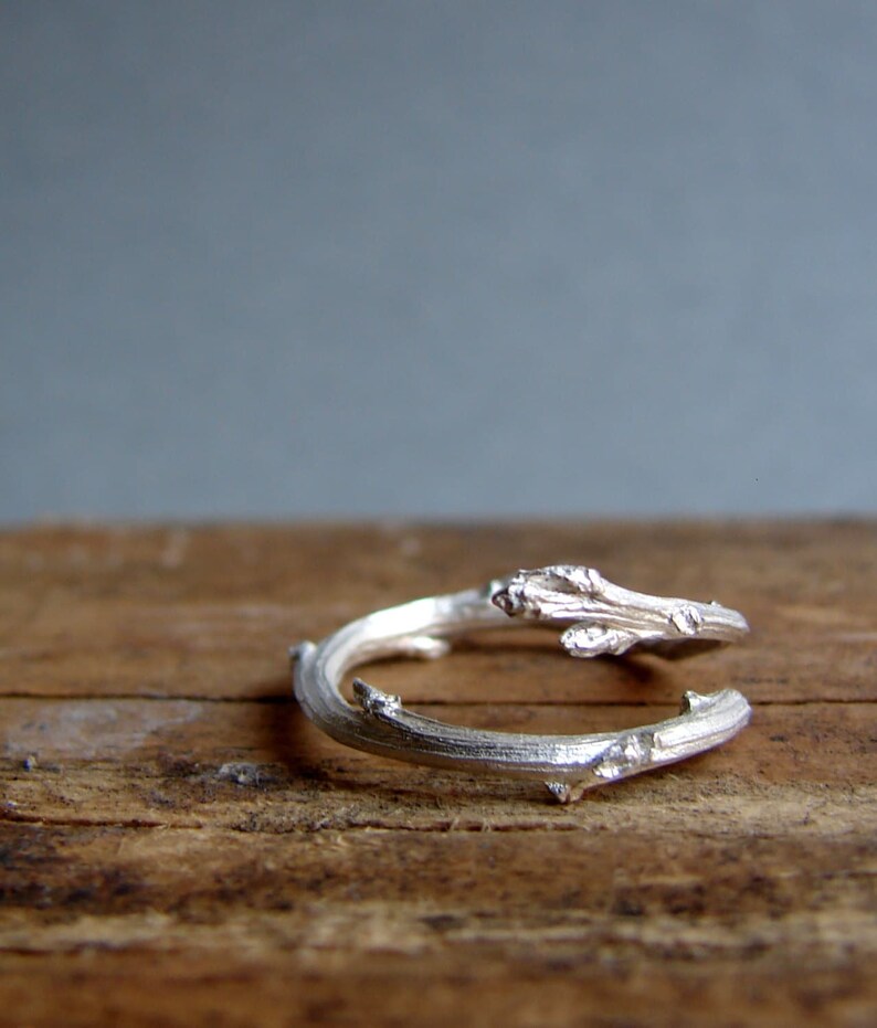 May include: A silver ring shaped like a twisted branch with small details. The ring is on a wooden surface.