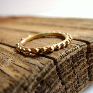 May include: A gold ring featuring a beaded design, set against a backdrop of aged wood. The ring's band is decorated with small, evenly spaced spherical beads. The wood has a rough, textured surface with visible grain.