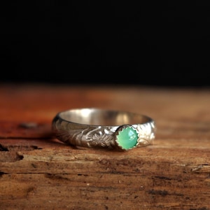 Könnte beinhalten: Ein silberner Ring mit floralem Muster und einem runden, hellgrünen Edelstein. Der Ring ist vor einem Holzuntergrund mit dunklem Hintergrund platziert, wodurch die filigranen Details des Metalls und die Farbe des Edelsteins hervorgehoben werden.