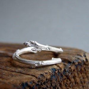 May include: A silver ring shaped like a twisted branch with small buds. The ring is on a piece of weathered wood.
