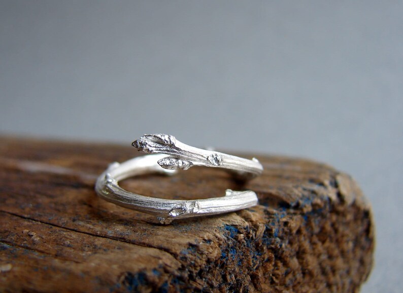 May include: A silver ring shaped like a twisted branch with small buds. The ring is on a piece of weathered wood.
