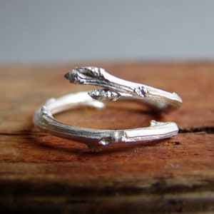 May include: A silver ring shaped like a twig with a small branch extending out. The ring is on a wooden surface.