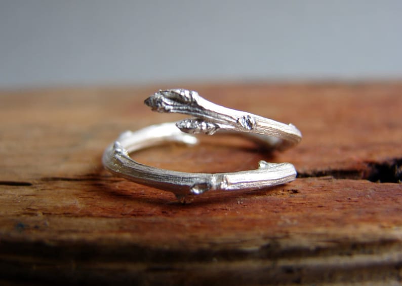 May include: A silver ring shaped like a twig with a small branch extending out. The ring is on a wooden surface.