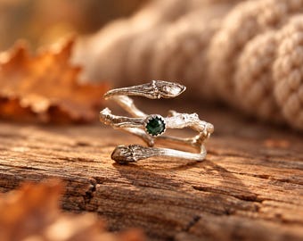 May include: A silver ring designed to resemble a textured tree branch that wraps around the finger. The ring features a single, small, round, dark green gemstone set securely in the center of the metal branch structure.