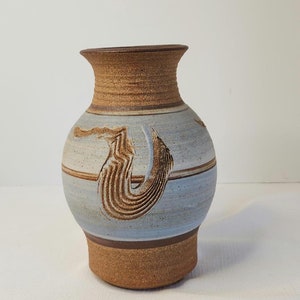 Può includere: Un vaso in ceramica fatto a mano con corpo azzurro e bordo e base marrone strutturati. Il vaso presenta un design astratto intagliato nei toni del marrone. Il vaso è alto circa 20 cm.