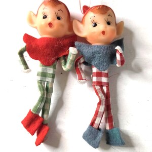 May include: Two elf ornaments with molded plastic faces, pointed ears, and painted features. One elf wears a red collar and green gingham suit with red boots. The other wears a blue collar and red gingham suit with blue boots. Both have hanging strings.