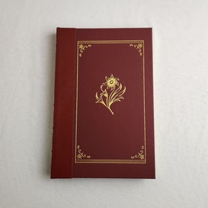 Pode incluir: Um diário encadernado em couro vermelho com um design floral dourado na capa.
